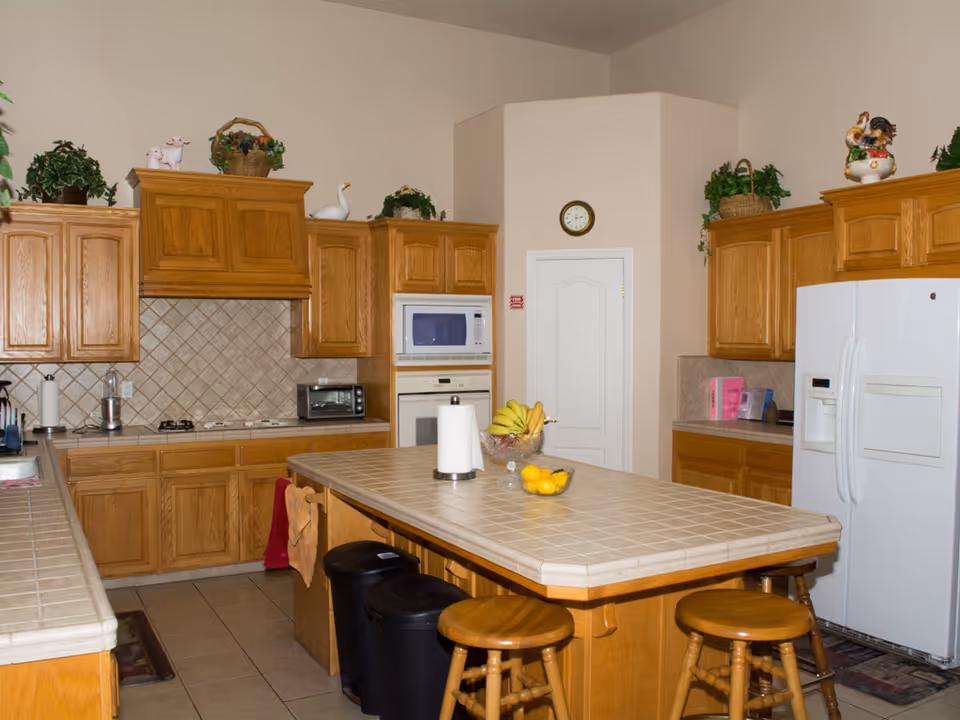 A spacious kitchen with wooden cabinets and a tiled countertop island in the center. The kitchen features a white refrigerator, built-in microwave and oven, a toaster oven, and a gas stove. There are decorative plants and figurines on top of the cabinets. The island has three wooden stools, a paper towel holder, a bowl of bananas, and a bowl of lemons. A clock is mounted on the wall above a white door in the background.