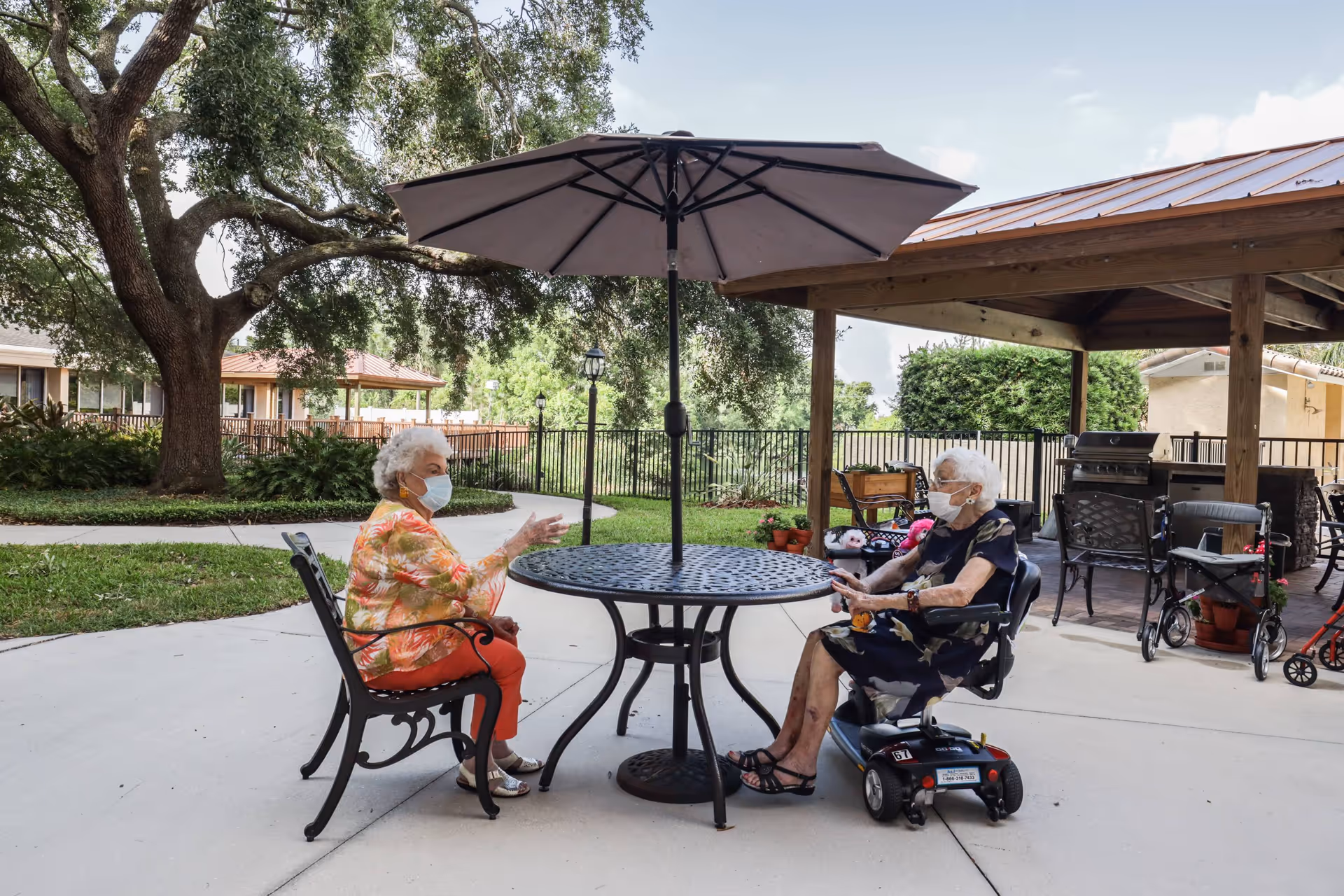 Two elderly women wearing face masks sitting outdoors at a round metal table with an umbrella. One woman is seated on a chair, and the other is on a mobility scooter. They are in a paved patio area with trees, plants, and a covered seating area with chairs and a grill in the background.