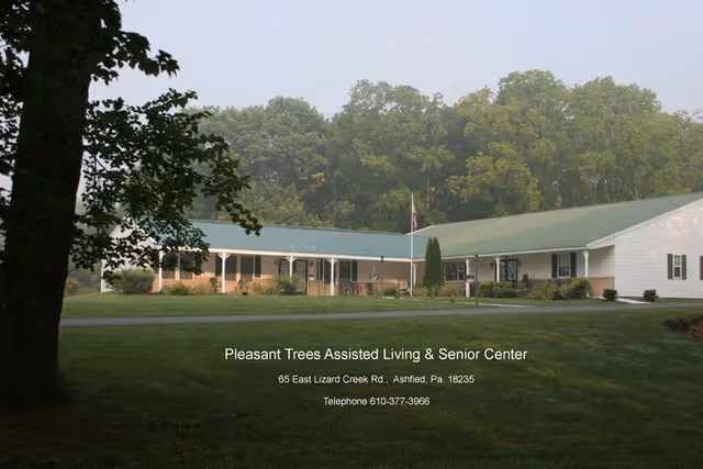 Exterior view of Pleasant Trees Assisted Living & Senior Center, a single-story building with a green roof surrounded by trees and a grassy lawn.