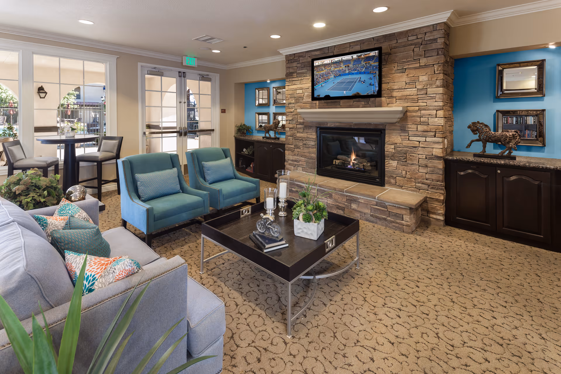 A cozy living room area in a senior living facility featuring a stone fireplace with a flat-screen TV mounted above it. The room has a beige patterned carpet, a gray sofa with colorful pillows, two teal armchairs, and a dark wood coffee table decorated with candles and a plant. There are large windows and glass doors leading to an outdoor patio with tables and chairs. The walls have blue accent sections with decorative mirrors and horse sculptures on dark wood cabinets.