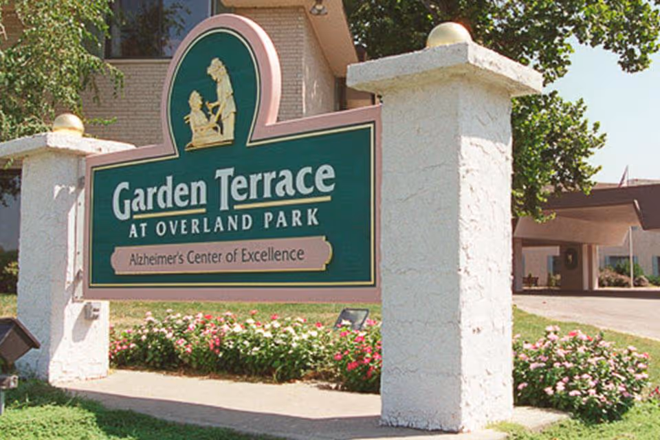 Entrance sign for Garden Terrace at Overland Park reading "Alzheimer's Center of Excellence" between stucco pillars with landscaping and the facility entrance behind it.