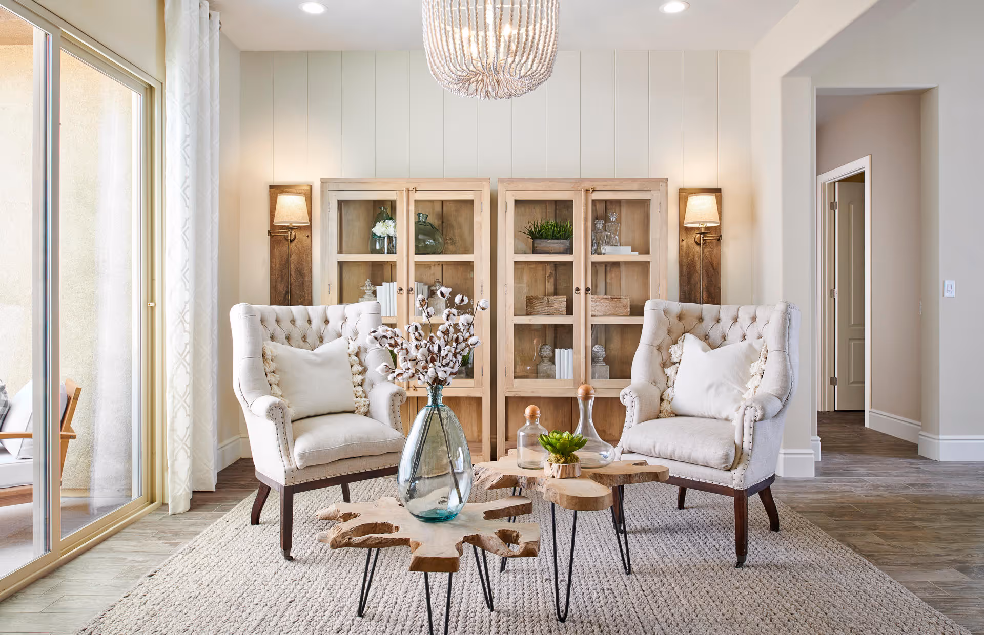 A bright and cozy living room area with two beige tufted armchairs facing each other, each with a white pillow. Between them are two wooden coffee tables with decorative items including a glass vase with cotton stems, a small plant, and glass bottles. Behind the chairs is a wooden cabinet with glass doors displaying books and decor. The room has light-colored walls, a textured rug, wall sconces with lamps, and a large window with sheer curtains letting in natural light.
