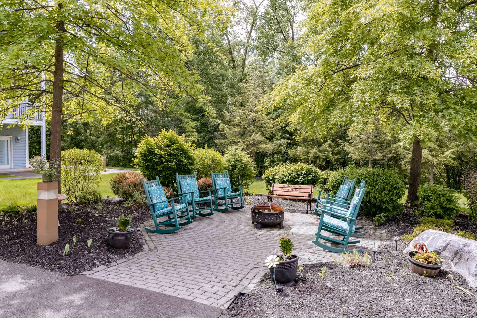 Outdoor seating area with six teal rocking chairs arranged in a circle around a fire pit on a paved patio. There is a wooden bench in the background with a memorial plaque, surrounded by green bushes and trees. The area is landscaped with mulch and potted plants, and a building with a balcony is partially visible on the left.