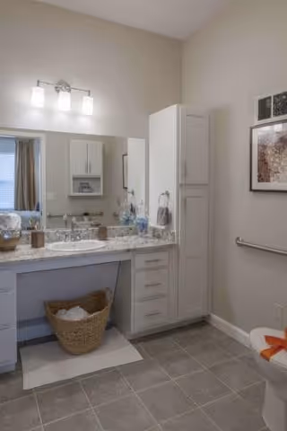 A clean and modern bathroom featuring a large mirror above a granite countertop with a sink, white cabinets and drawers, a wicker basket with towels underneath the counter, a toilet with an orange towel on the seat, a grab bar on the wall, and framed artwork hanging above the grab bar.