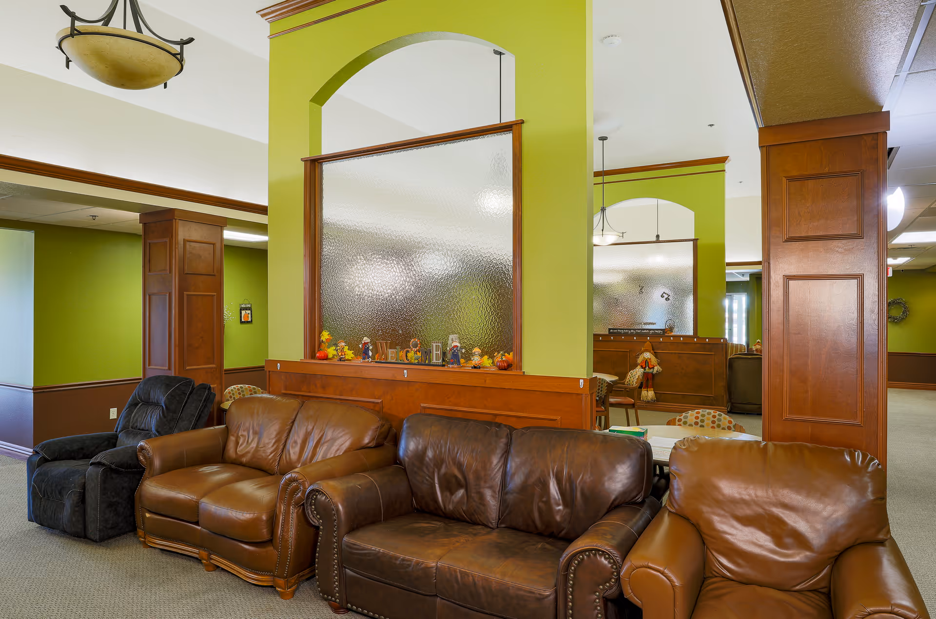 A cozy seating area in a senior living facility with three leather sofas and one recliner chair arranged along a green and brown wall with wooden trim. The space is decorated with small fall-themed figurines on a wooden ledge beneath a frosted glass partition. The room has a warm and inviting atmosphere with soft lighting and a carpeted floor.