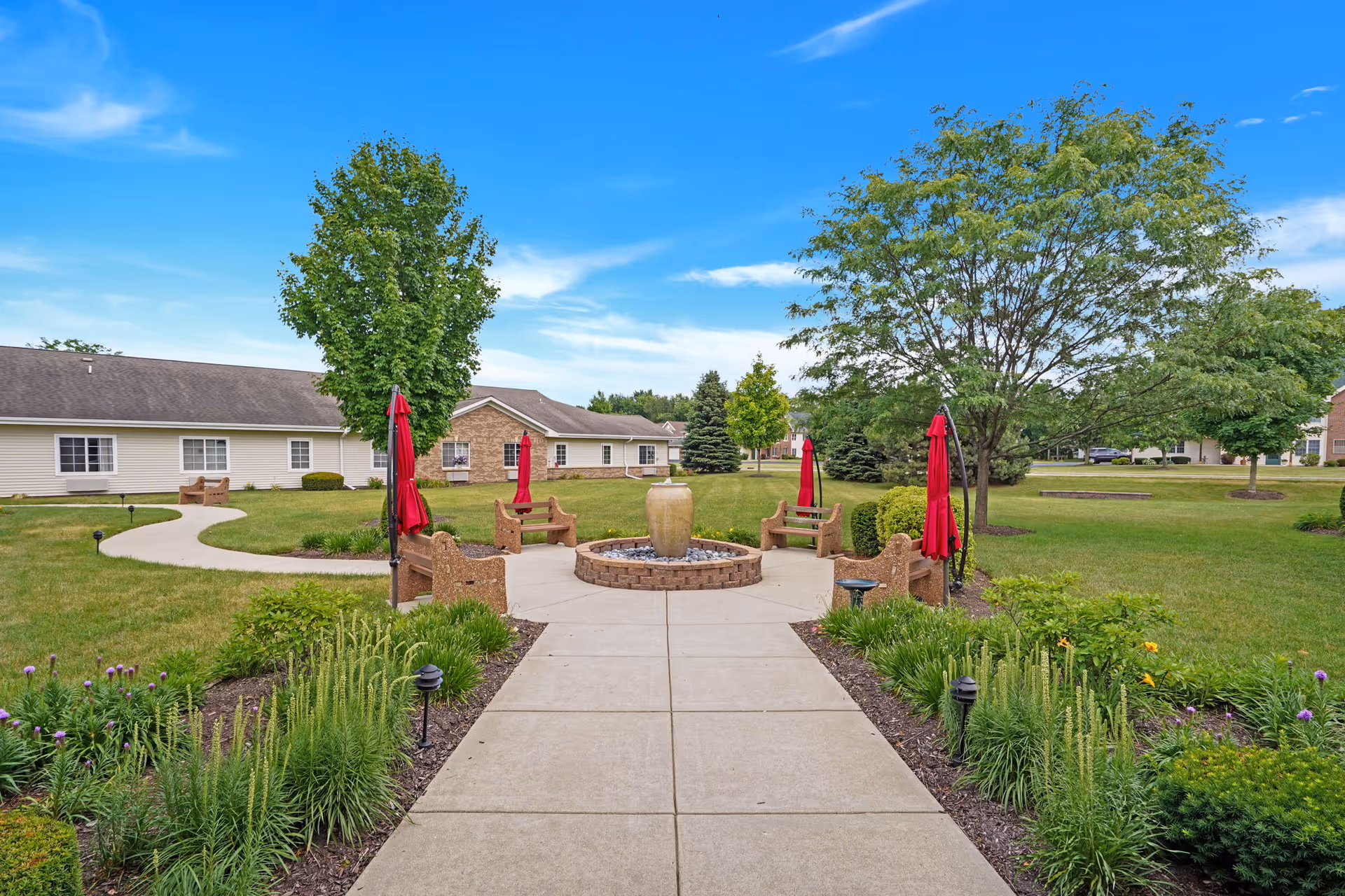 Paved walkway leading to a circular fountain with benches and red umbrellas in a grassy courtyard in front of single-story senior living buildings.