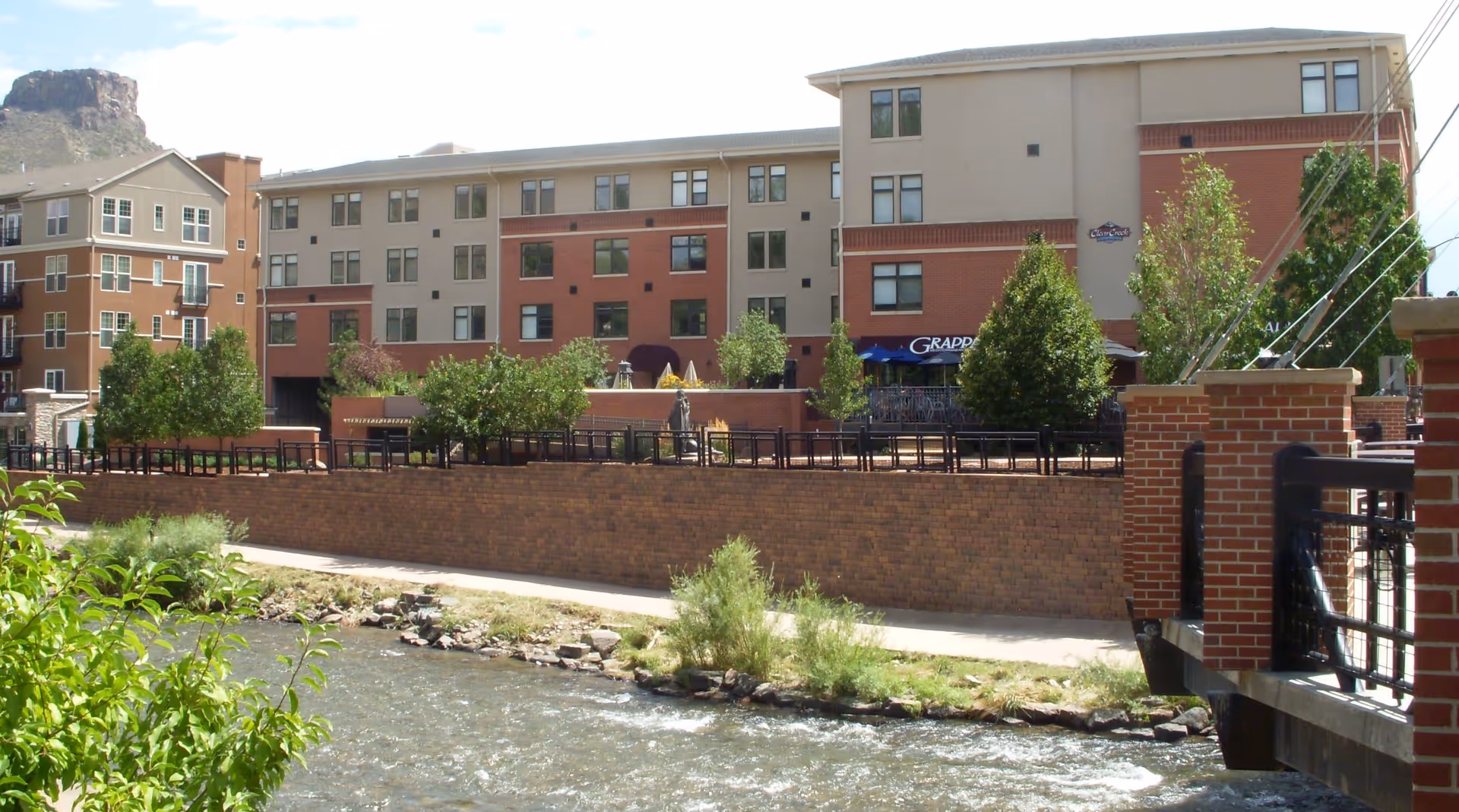 Exterior view of a multi-story brick and stucco building by a river with a riverside walkway, trees, and a small pedestrian bridge.