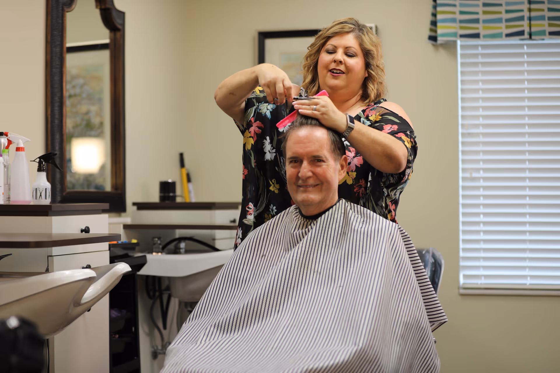 A woman is cutting a man's hair in a salon setting. The man is seated and covered with a striped barber cape, smiling at the camera. The woman is standing behind him, holding hair clippers and a pink comb. The room has a large mirror, a window with blinds, and various hair care products on the counter.
