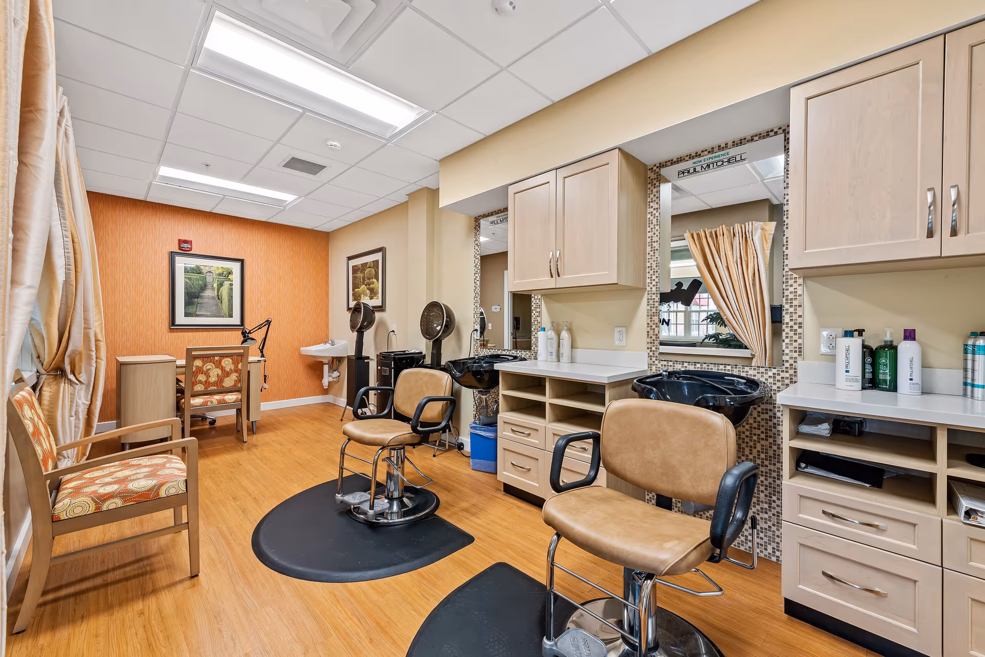Interior view of a salon area in a senior living facility with two salon chairs in front of mirrors and sinks, a small desk with a chair, a patterned armchair, and framed artwork on the walls. The room has wood flooring and light-colored cabinetry.