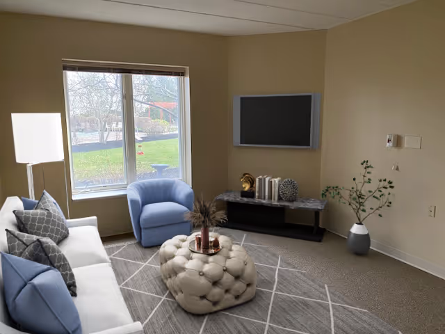 A cozy living room with a large window showing an outdoor garden. The room features a white sofa with blue and gray pillows, a blue armchair, a tufted beige ottoman with a decorative tray, a modern floor lamp, a flat-screen TV mounted on the wall above a console table with books and decorative items, and a potted plant in the corner.