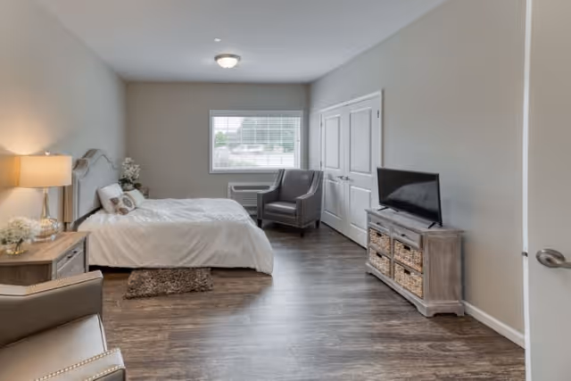 A bright and spacious bedroom with a large bed covered in white bedding, a bedside table with a lamp and flowers, two gray armchairs near a window with blinds, a wooden dresser with a flat-screen TV on top, and wood flooring throughout.