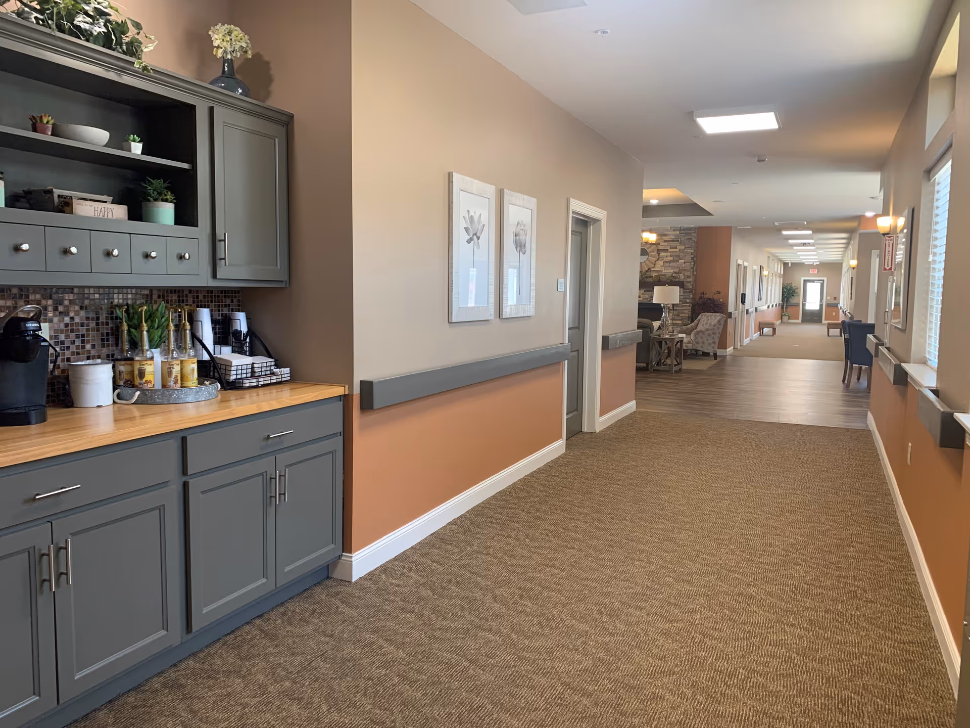 A well-lit hallway in a senior living facility with beige and orange walls, carpeted floor, and a wooden countertop with cabinets on the left side. The countertop holds a coffee maker, cups, and decorative plants. The hallway extends into a common area with seating and a stone accent wall with a clock. Artwork is hung on the walls, and windows on the right side allow natural light.