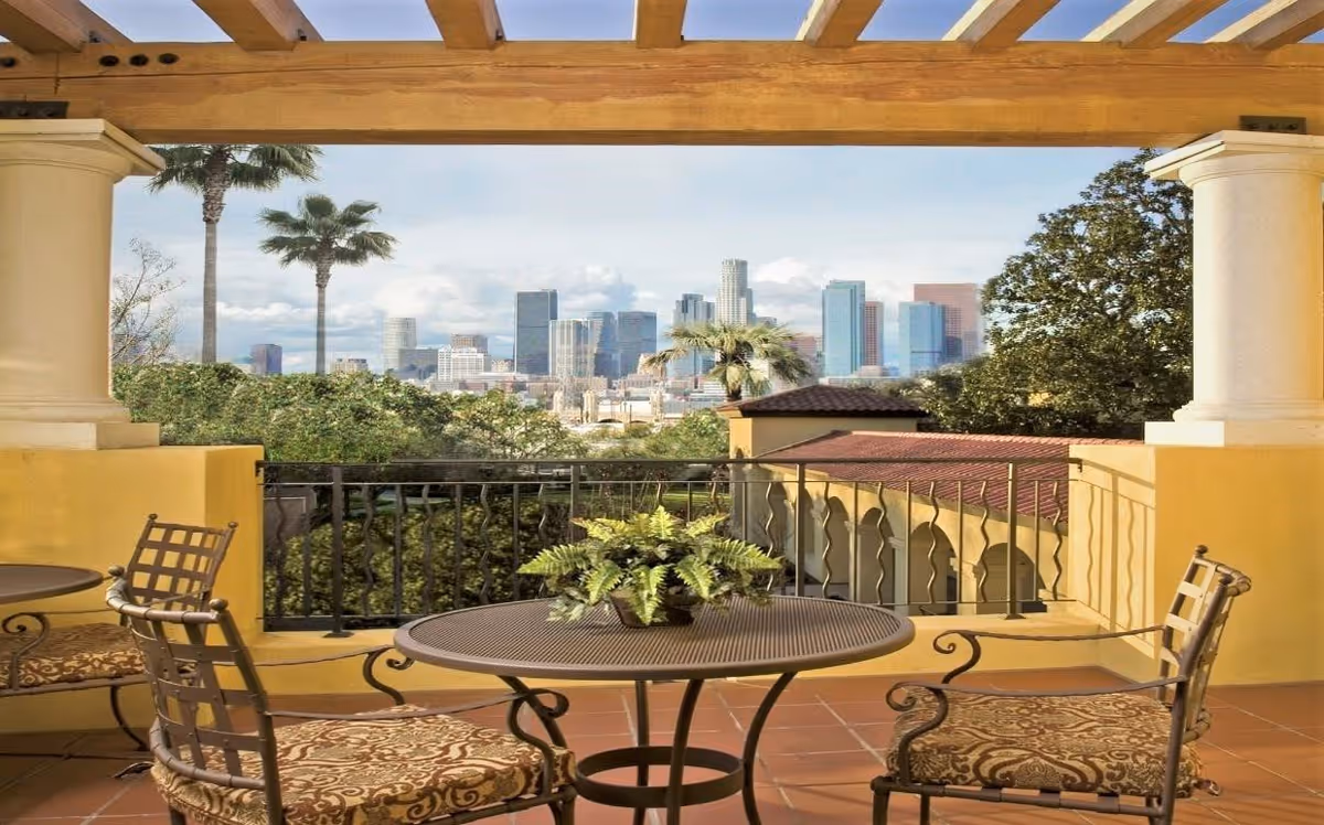Outdoor patio area with a round metal table and four cushioned chairs under a wooden pergola, overlooking greenery and a city skyline in the background.