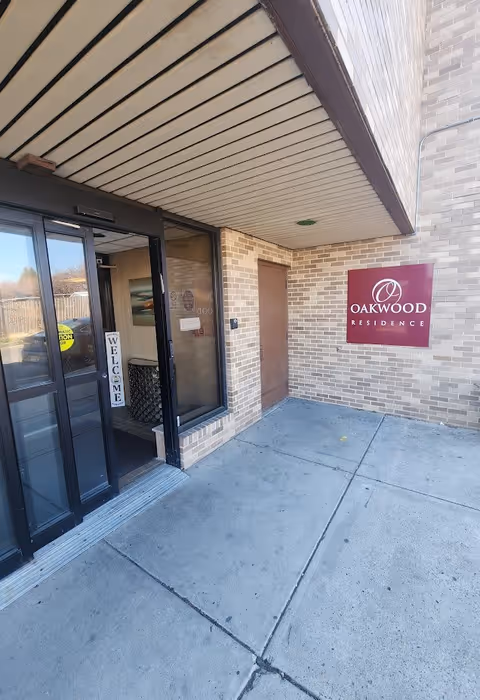 Entrance area of Oakwood Residence showing a glass door with a welcome sign inside, a brick wall with a maroon sign displaying the Oakwood Residence logo, and a concrete walkway.