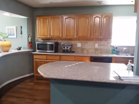 Interior view of a kitchen with wooden cabinets, a microwave, a coffee maker, a dishwasher, and a sink under a window. The kitchen has a curved countertop with a granite surface and green walls.