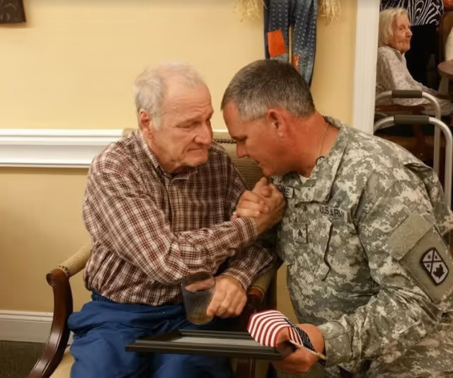 An elderly man sitting in a chair shakes hands with a man in a U.S. Army uniform who is kneeling beside him. The elderly man holds a glass and a framed item with a folded American flag. In the background, another elderly person is seated in a wheelchair.