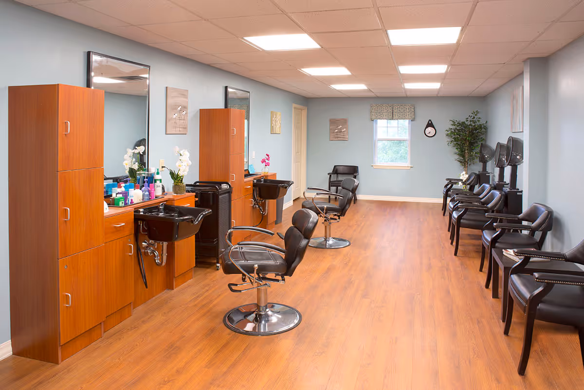 Interior of a hair salon with wooden flooring, light blue walls, and ceiling lights. There are two wooden stations with black sinks and mirrors, each accompanied by a black salon chair. On the right side, there are multiple black chairs lined up against the wall, some with hair drying machines. A window with a patterned valance and a wall clock are visible at the far end, along with a potted plant.