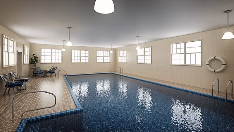 Indoor swimming pool with blue water surrounded by beige tiled walls and floor. Several windows allow natural light to enter the room. There are metal handrails for pool entry and exit, a life preserver mounted on the wall, and seating areas with chairs and a small table near a potted plant.