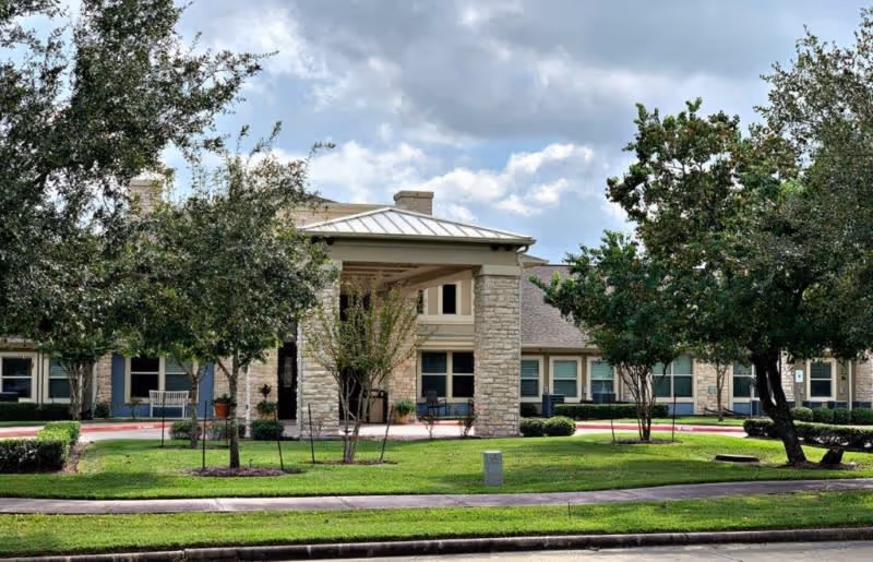 Exterior view of a senior living facility building with a covered entrance supported by stone pillars, surrounded by green grass, trees, and shrubs under a cloudy sky.