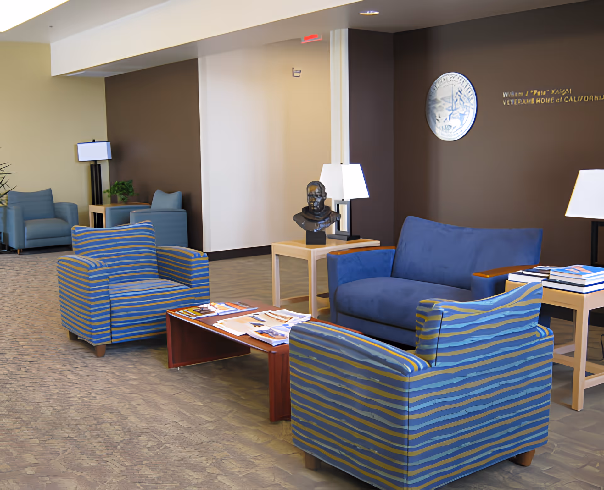 A seating area in a facility with two blue and yellow striped armchairs, a blue loveseat, a wooden coffee table with magazines, side tables with lamps, a bust sculpture, and a brown accent wall with a round emblem and partial gold lettering.