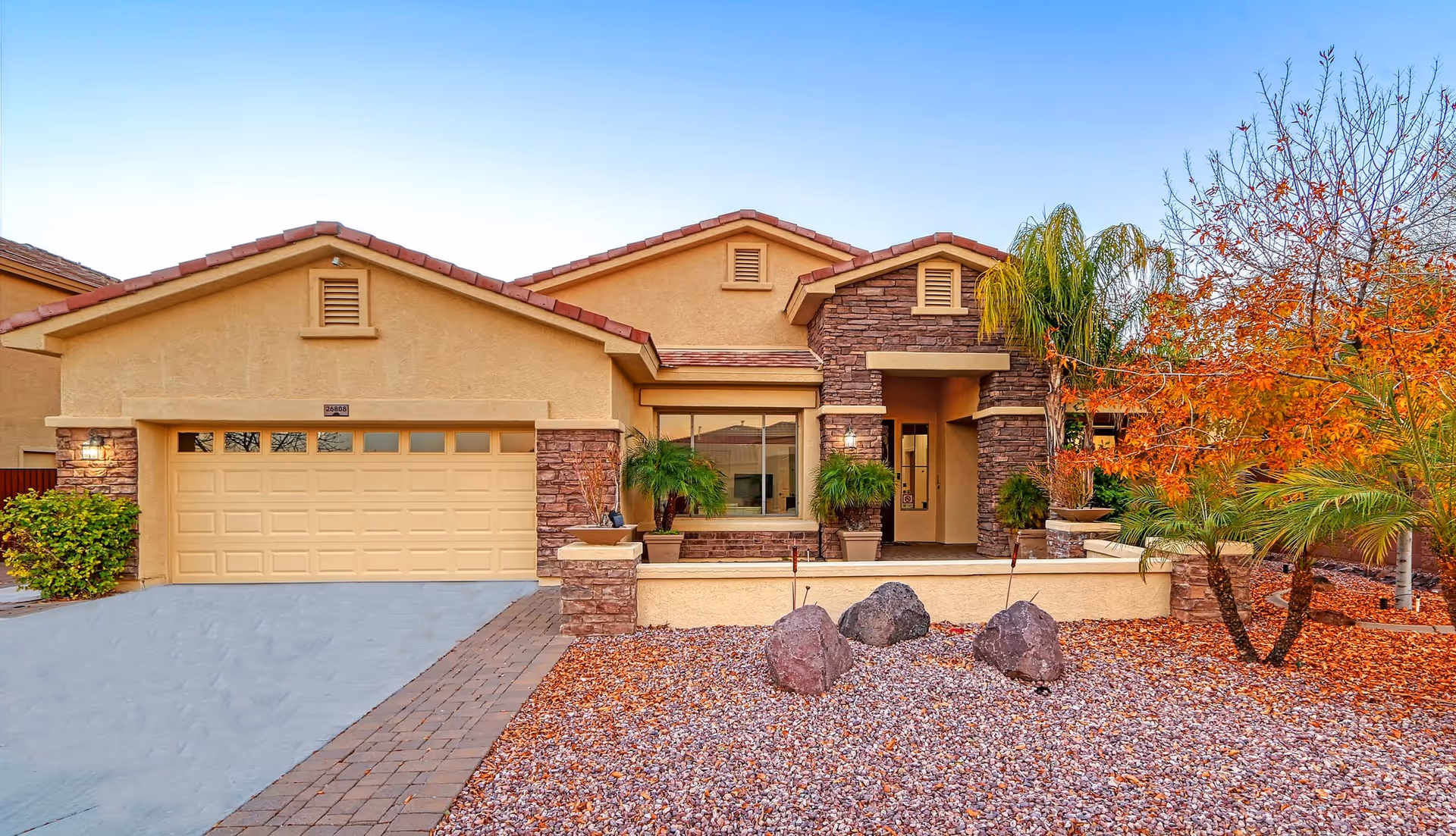 Front exterior view of a single-story house with a two-car garage, stone and stucco facade, a small porch with potted plants, and a landscaped front yard with rocks, palm trees, and autumn-colored foliage.