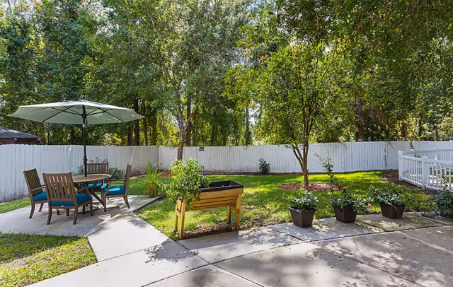 Sunny fenced backyard patio with a table and umbrella, chairs, potted plants and a raised planter among trees.