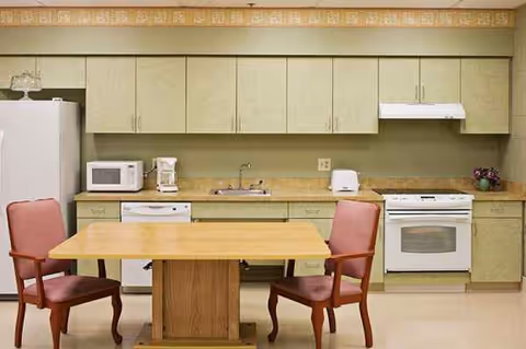 Communal kitchen with beige cabinets, a wooden table, and pink upholstered chairs.
