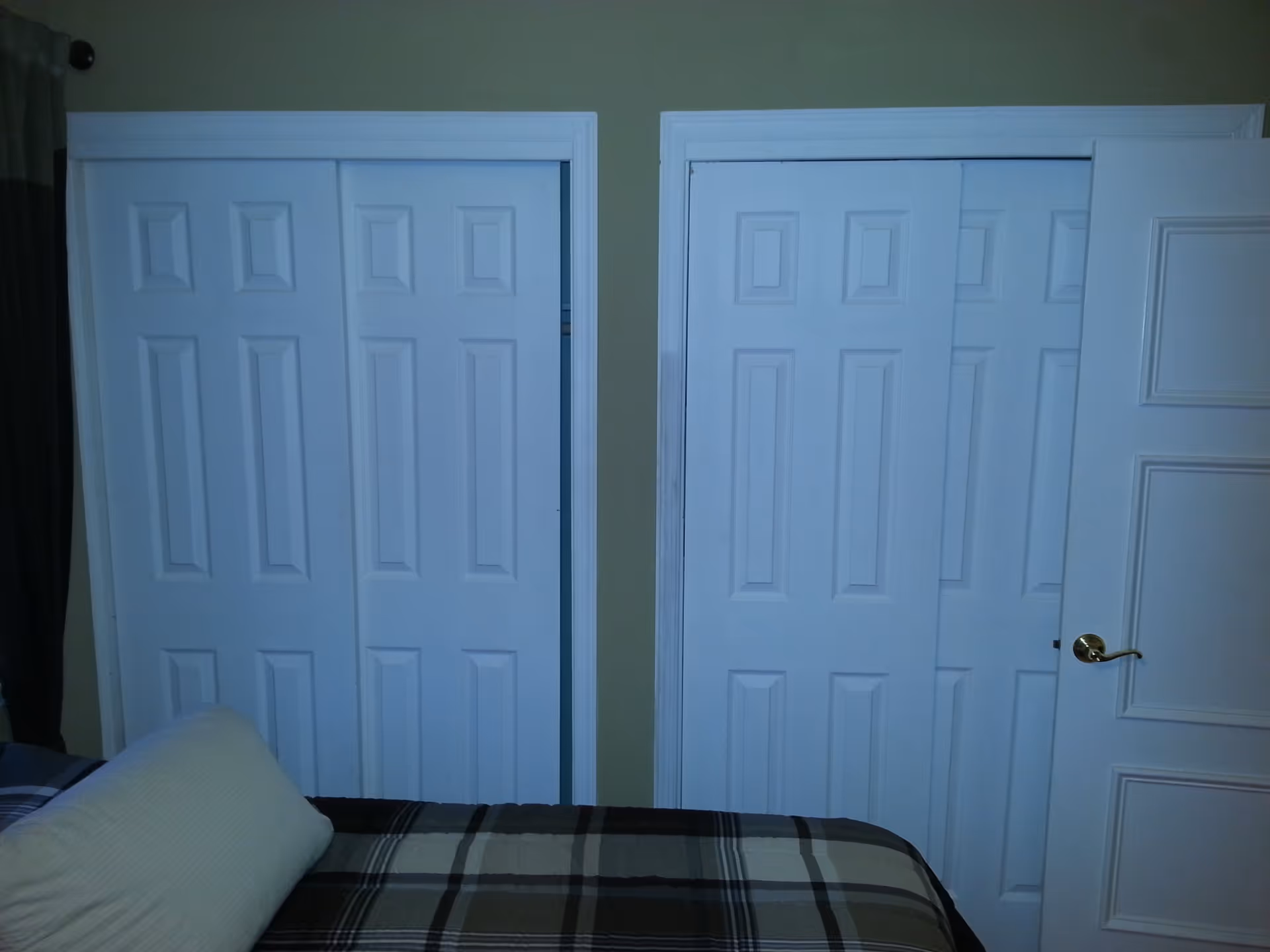 Interior view of a bedroom showing two white sliding closet doors side by side on a beige wall, with part of a bed covered in a plaid blanket and a light-colored pillow visible in the foreground.