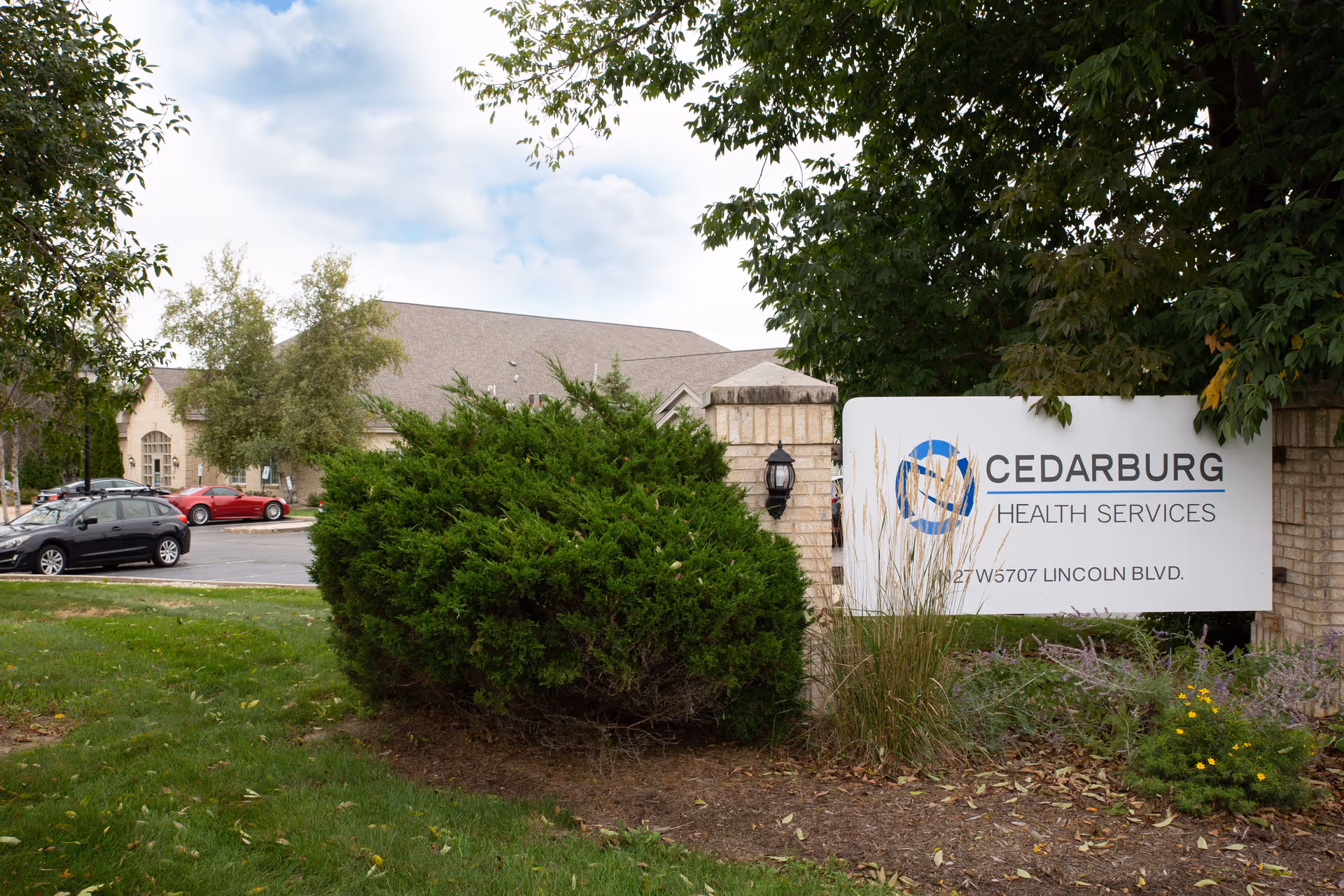 Outdoor view of Cedarburg Health Services facility sign surrounded by greenery and trees, with a parking lot and building visible in the background under a partly cloudy sky.