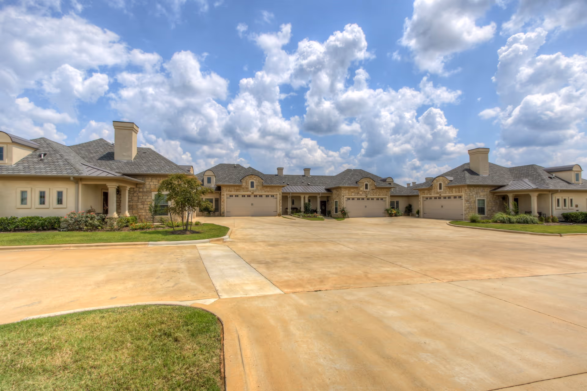 Wide exterior view of a senior living facility with multiple attached units featuring garages, beige walls, stone accents, and a large paved driveway under a partly cloudy sky.