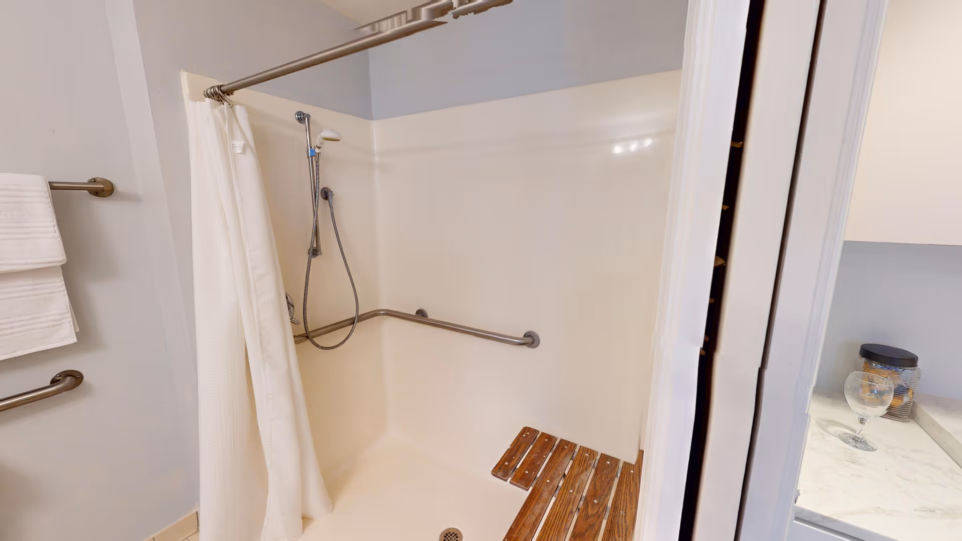 A bathroom shower area with a white shower curtain, a handheld showerhead, and a wooden fold-down shower seat. There are grab bars installed on the walls for support, and a towel rack with white towels is visible on the left side.