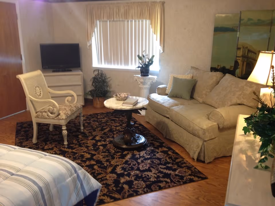 A cozy living room in an assisted living facility featuring a beige sofa with patterned cushions, a white armchair with a floral cushion, a round marble-top coffee table with books and a decorative bowl, a patterned area rug, a flat-screen TV on a white dresser, a potted plant, and a window with vertical blinds and a valance.