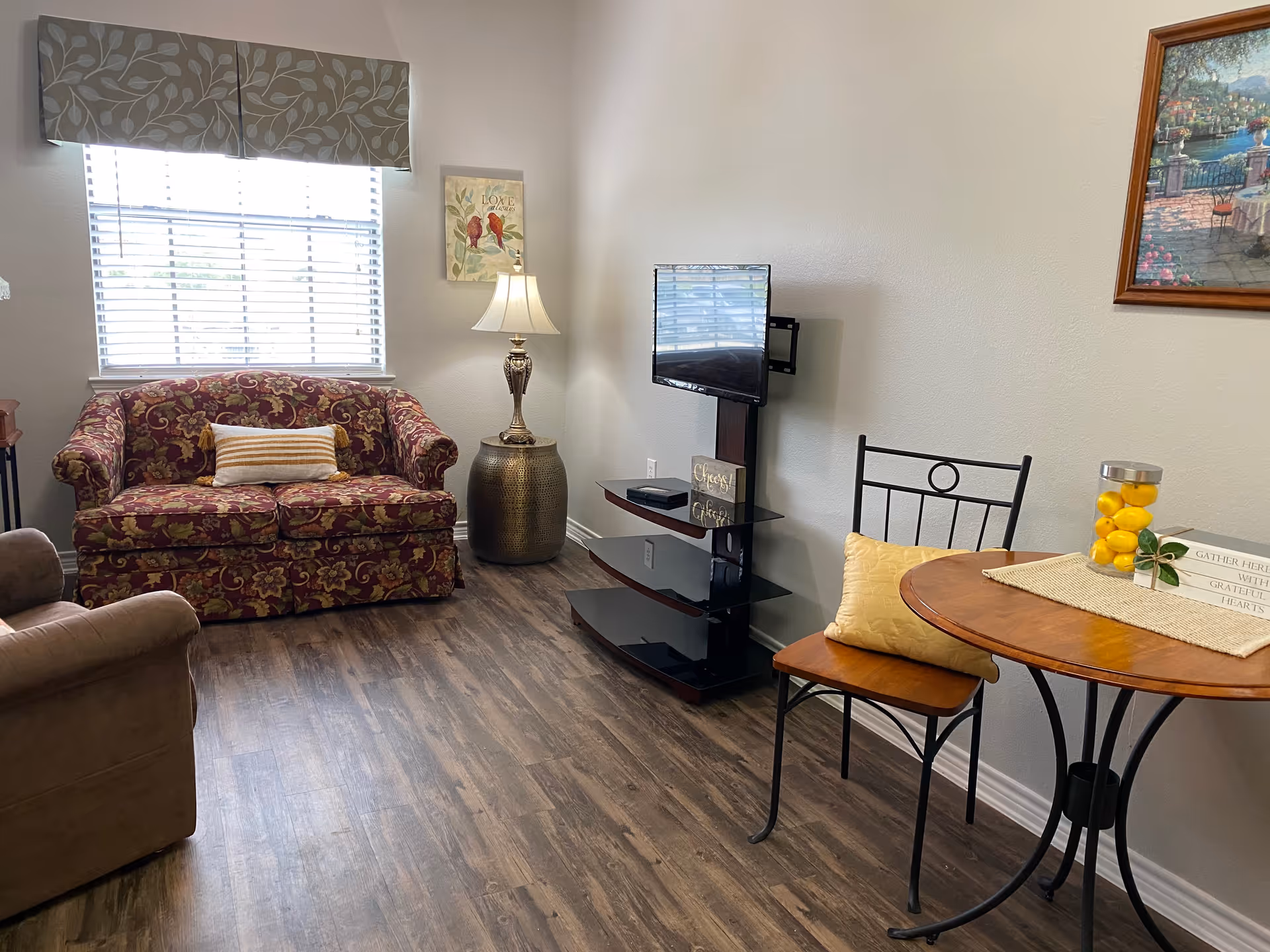 A cozy living room with a floral patterned loveseat, a brown armchair, a small round wooden table with a yellow cushion on a metal chair, a TV mounted on a black stand, a decorative lamp on a round side table, and wall art. The room has wood flooring and a window with blinds and a valance.