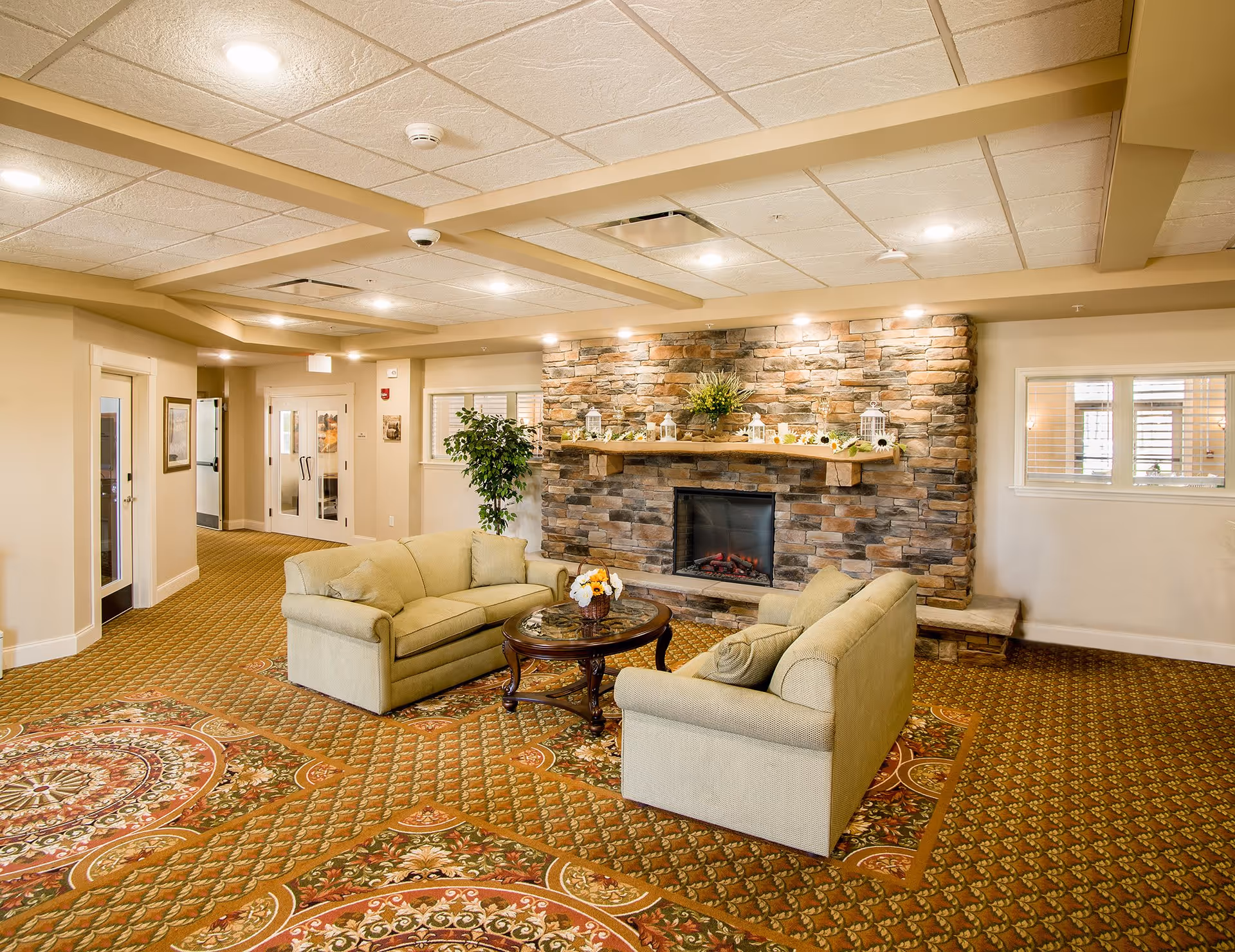 A cozy common area in a senior living facility featuring two beige sofas arranged around a round wooden coffee table with a floral centerpiece. Behind the seating area is a stone fireplace decorated with small lanterns and flowers on the mantel. The room has a patterned carpet, beige walls, and a ceiling with recessed lighting and beams. There are windows and doors visible in the background.