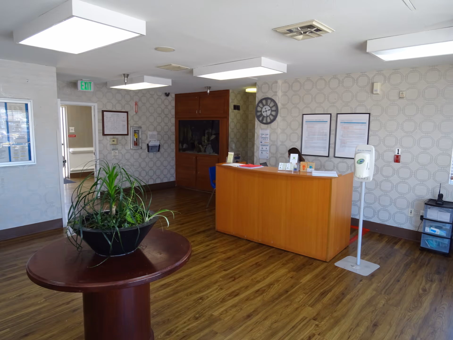 Reception area inside Rosecrans Care Center featuring a wooden reception desk with informational pamphlets, a hand sanitizer stand, a round table with a large potted plant, wood flooring, patterned wallpaper, a wall clock, and framed notices on the wall.