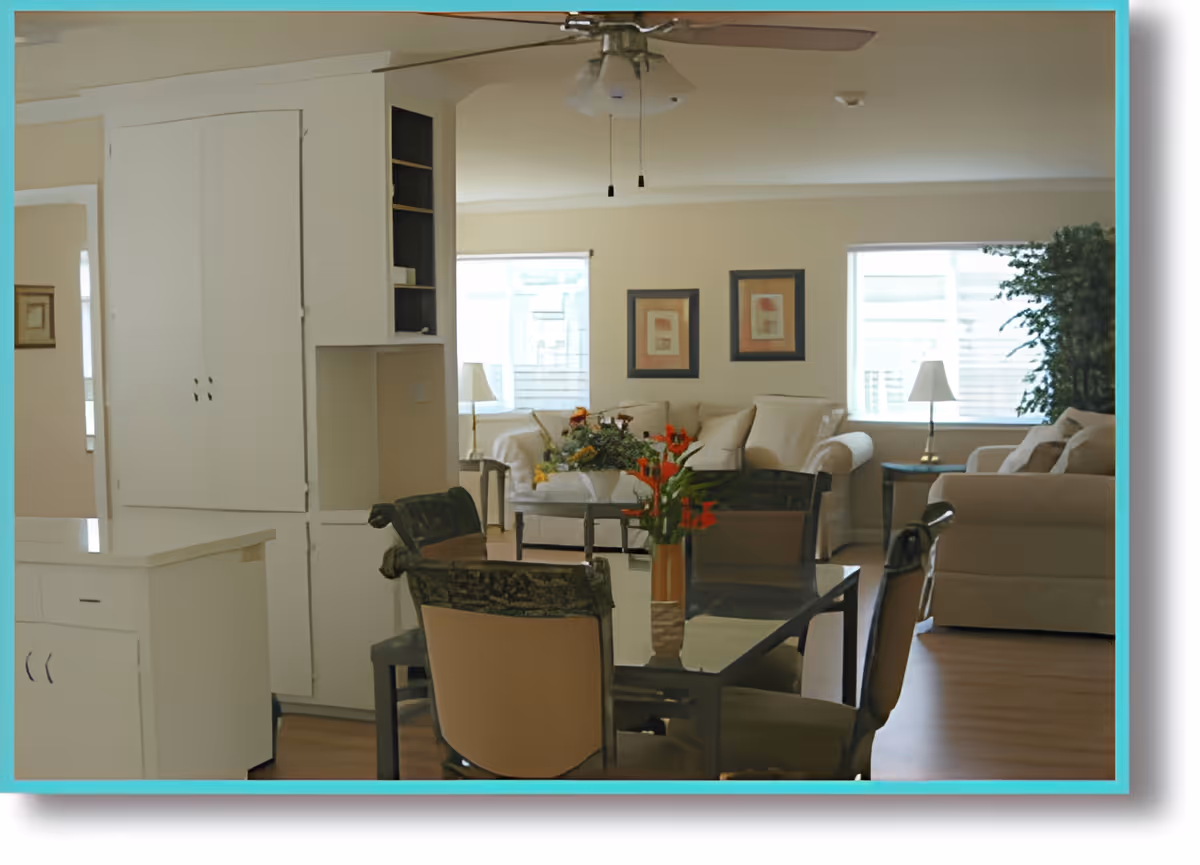 A bright living room and dining area with beige sofas, a glass dining table with chairs, a ceiling fan, two framed artworks on the wall, two windows letting in natural light, and a potted plant in the corner.