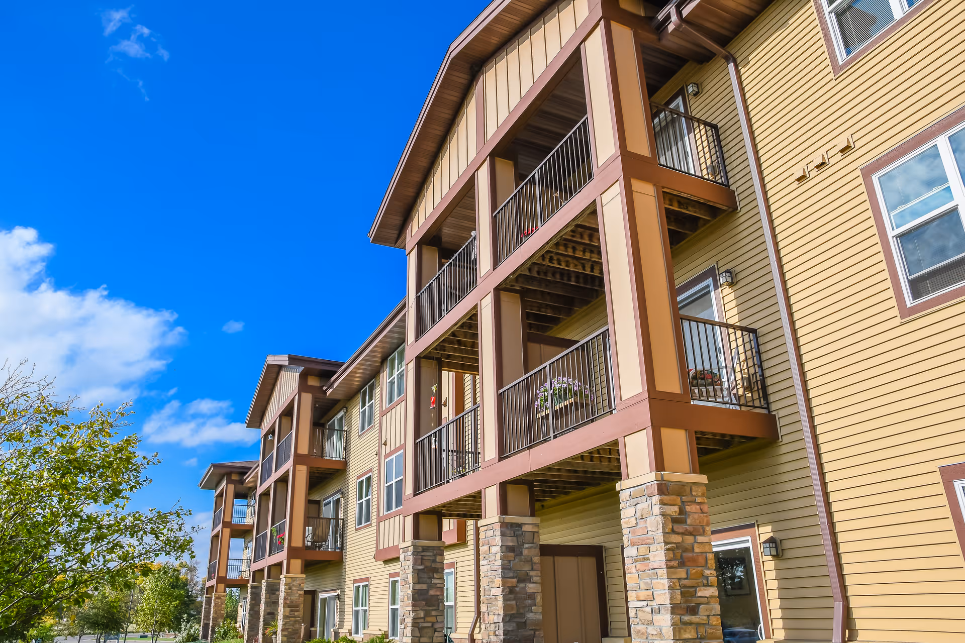 Exterior view of a multi-story residential building with beige siding, stone pillars, and balconies under a clear blue sky.