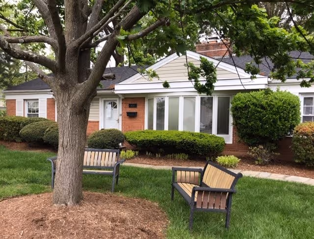 Exterior view of a single-story brick building with white trim and a large tree in the foreground. Two wooden benches with metal frames are placed on the grass near the tree. Shrubs and landscaping surround the building.