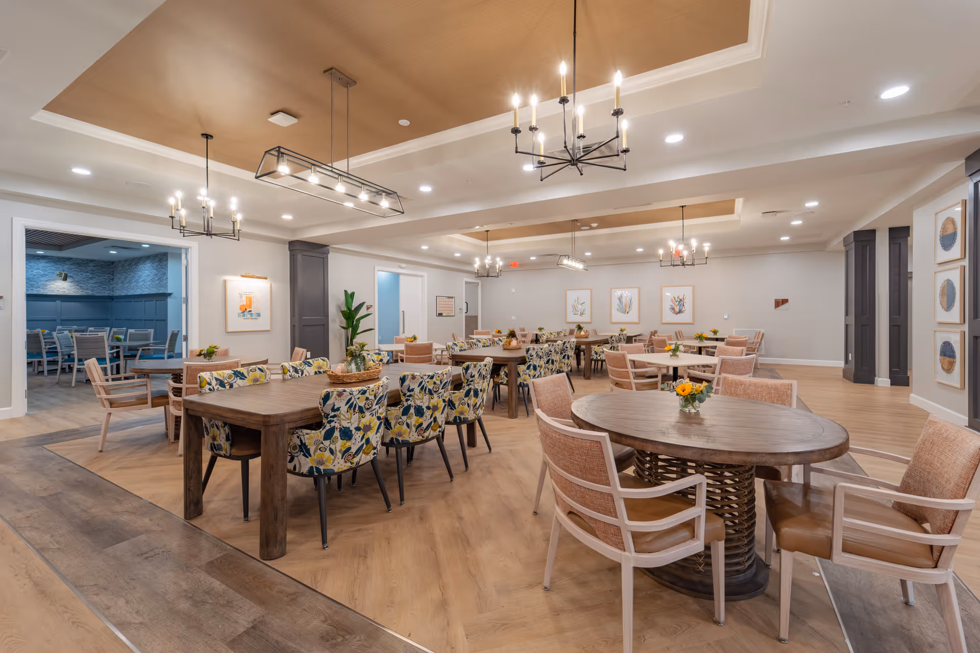 A spacious and well-lit dining room in a senior living facility with multiple wooden tables and chairs. Some chairs have floral upholstery while others have a solid peach color. The room features modern chandeliers, light wood flooring, and framed artwork on the walls. There is an adjoining room visible with additional seating and blue walls.
