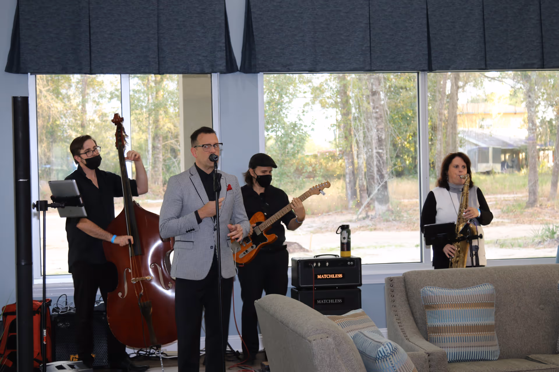 A small band performs in a senior living lounge—singer at a microphone with upright bass, electric guitar, and saxophone in front of large windows and couches.