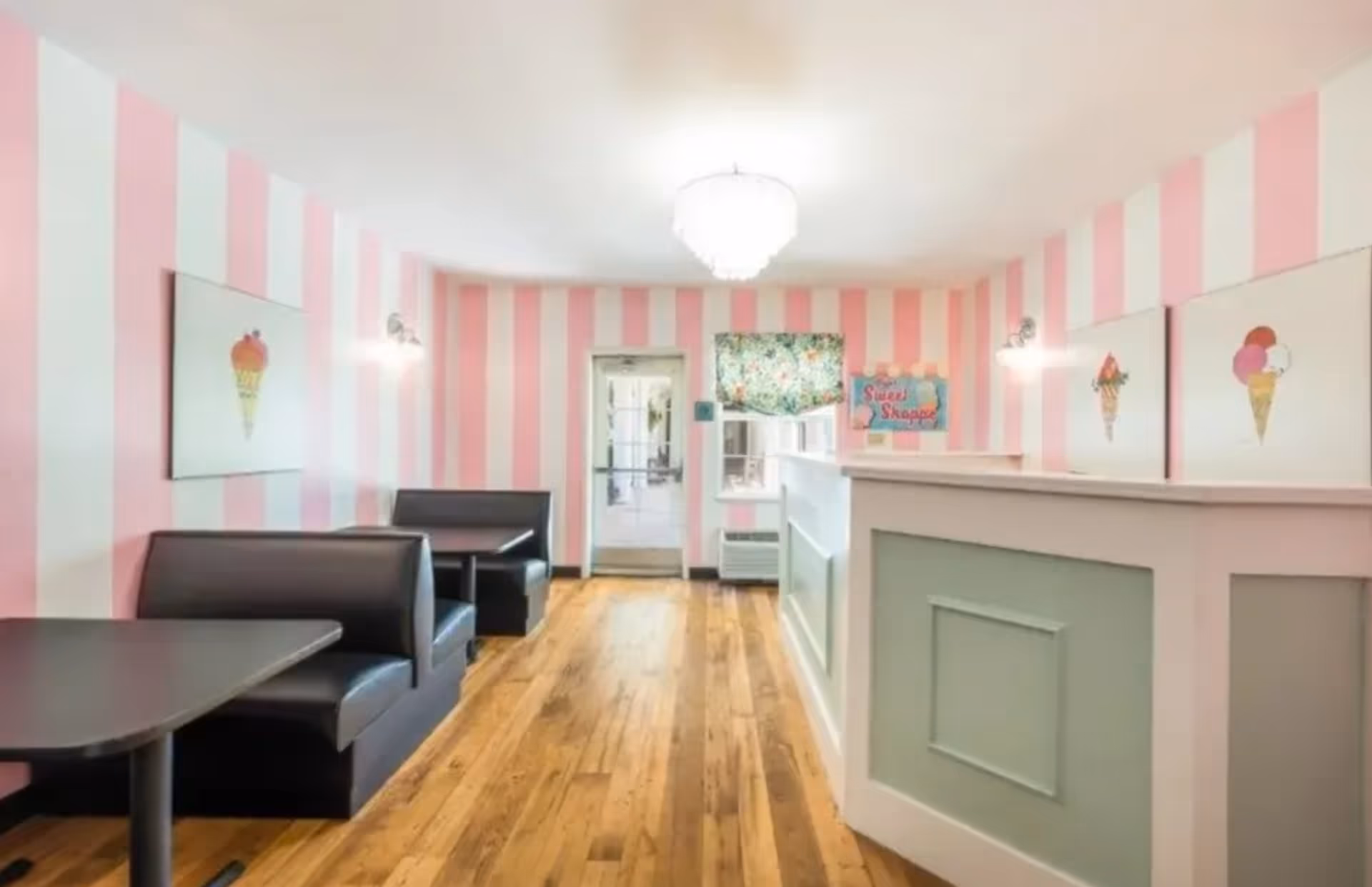 Interior of a small dining area with pink and white striped walls, wooden flooring, black booth seating with tables on the left, and a white and green counter on the right. The walls are decorated with pictures of ice cream cones and a sign that reads 'Sweet Shoppe'. A chandelier hangs from the ceiling.