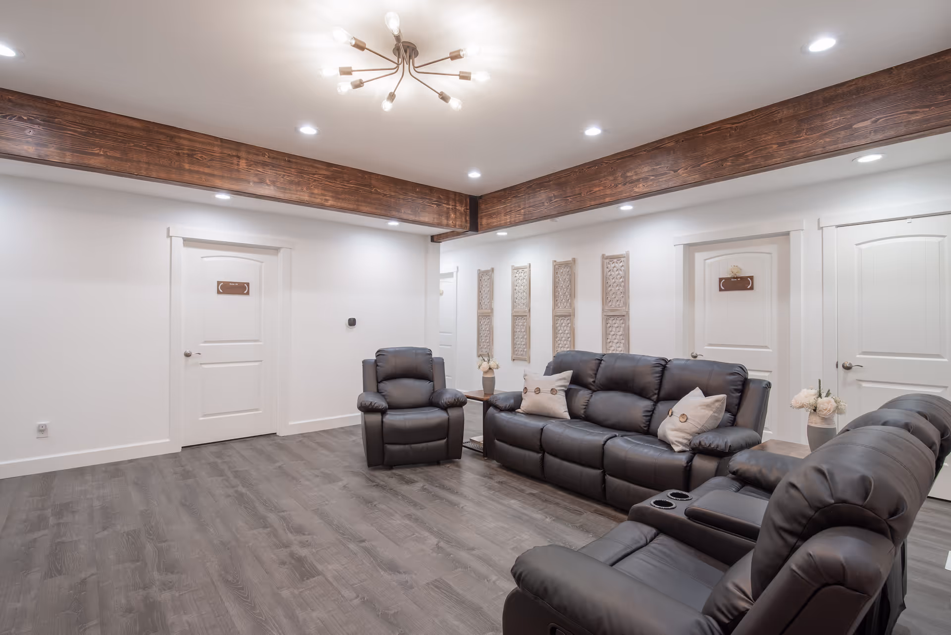 A cozy senior living facility common area with dark leather recliner chairs and a matching sofa, light gray walls, wooden ceiling beams, recessed lighting, and decorative wall panels. There are three white doors along the walls and a modern chandelier hanging from the ceiling.