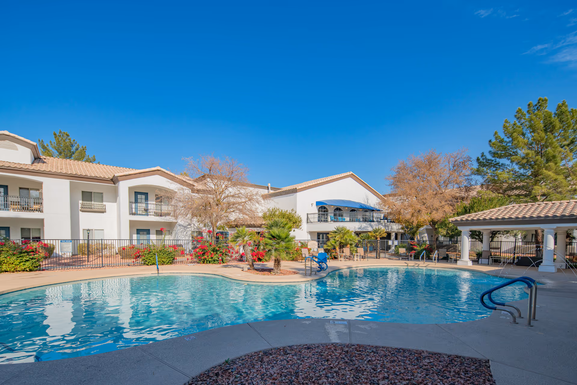 Outdoor swimming pool and poolside seating in front of two-story white Mediterranean-style senior living buildings under a clear blue sky.