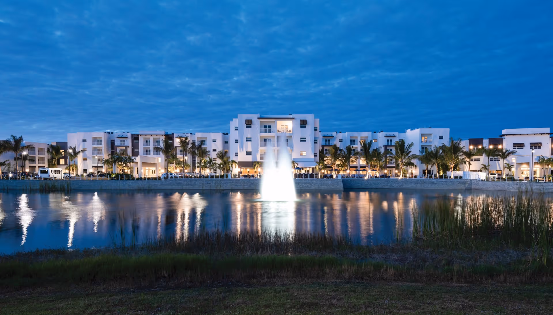 Evening view of a large, modern senior living facility named Amavida at Lakes Park with white buildings, palm trees, and a central water fountain illuminated in a lake in front of the building.