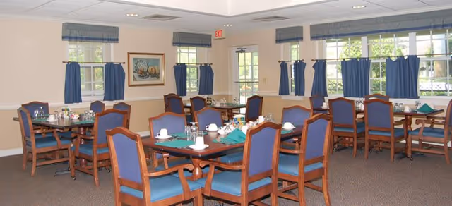 A dining room with multiple wooden tables and chairs with blue cushions. Each table is set with white cups, glasses, and green napkins. The room has several windows with blue curtains and valances, beige walls, and a framed painting on one wall.
