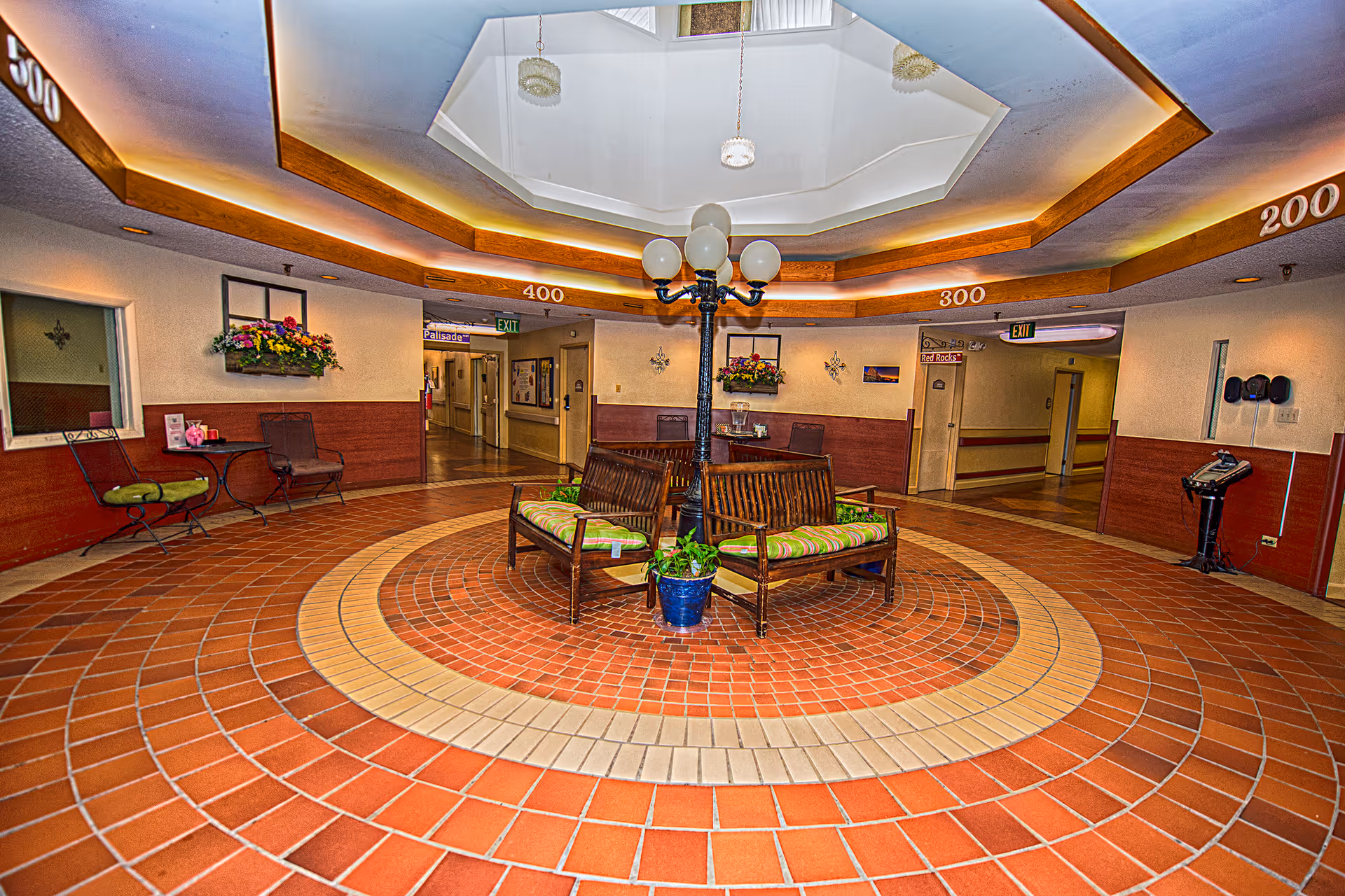 A circular lobby area with a tiled floor featuring concentric circles in shades of red and beige. In the center, there are two wooden benches with green striped cushions and a black lamp post with five globe lights. The ceiling above has a recessed octagonal design with wood trim and soft lighting. The walls have wooden paneling on the lower half and light-colored paint above, decorated with flower arrangements and framed pictures. Hallways labeled with room numbers 200, 300, and 400 lead off the lobby, with exit signs visible.