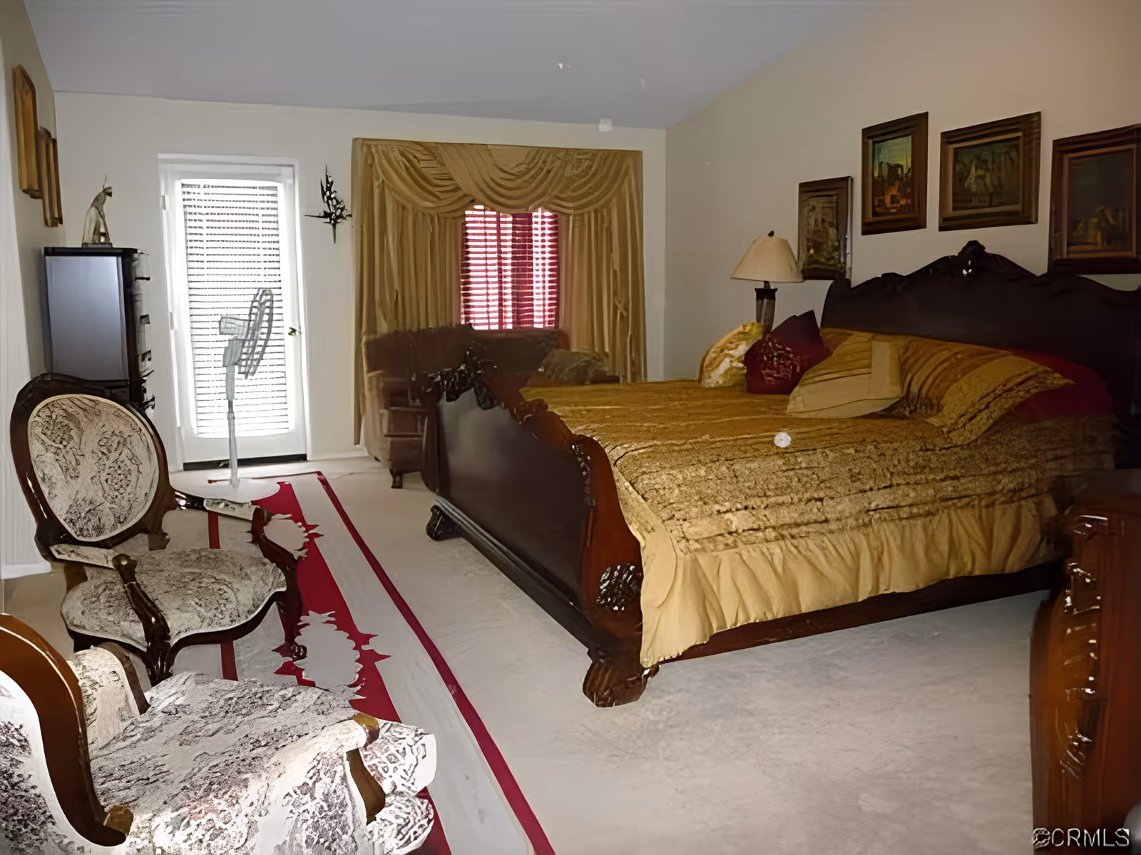 A bedroom with a large wooden bed covered with a gold bedspread and multiple pillows. There are two upholstered chairs with wooden frames and patterned fabric, a standing fan near a glass door with blinds, a wooden dresser, and framed artwork on the walls. The room has beige walls and carpeted floor with a red and white rug.