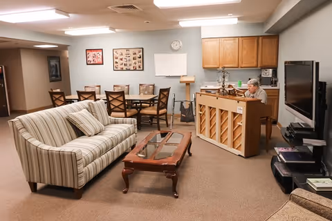 A cozy common area in a senior living facility featuring a striped sofa with a matching pillow, a wooden coffee table with glass inserts, a dining table with six chairs, and a piano being played by an elderly person. The room has beige carpeting, light-colored walls, wooden cabinets, a wall-mounted TV, and framed photos on the wall.