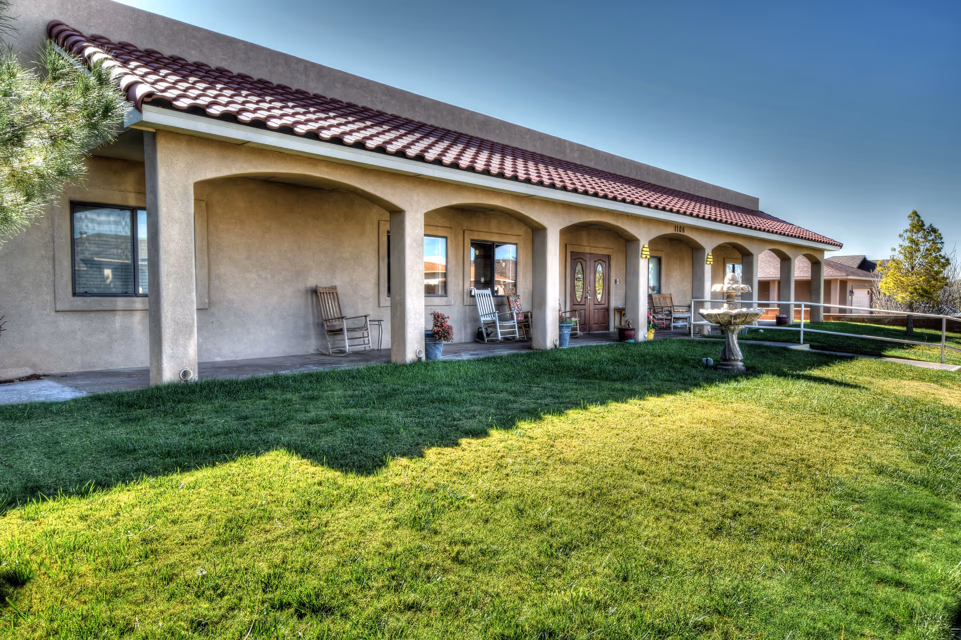 Exterior view of a single-story building with a red tile roof and beige stucco walls. The front porch has several rocking chairs and potted plants, with a green lawn and a decorative fountain in the foreground under a clear blue sky.