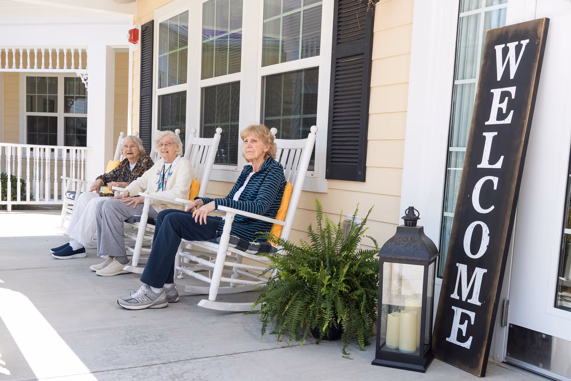 Three elderly women sitting on white rocking chairs on a porch outside a building with large windows and black shutters. There is a large vertical wooden sign that says 'WELCOME' and a black lantern with candles next to a green potted fern on the porch floor.